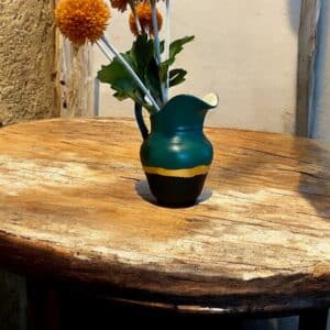 Petit pichet relooké en vert bouteille et bleu marine avec un liseré en feuille d’or, posé sur une table en bois rustique avec un bouquet de fleurs orange