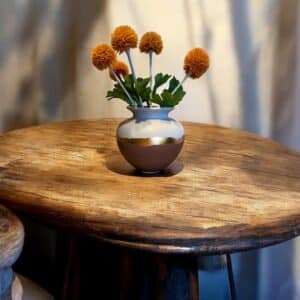 Vase rond rose et blanc avec un liseré en feuille d’or posé sur une table en bois rustique, avec des fleurs orange