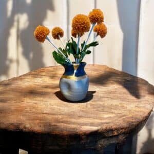 Petit vase bleu marine et gris avec un liseré d’or posé sur une table en bois rustique, avec un bouquet de fleurs orange