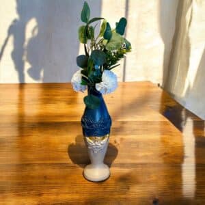 Vase haut et fin beige et bleu avec moulure et liseré en feuille d’or posé sur une table en bois rustique, avec des feuilles d’eucalyptus