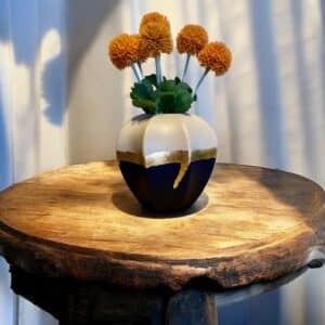 Vase beige et bleu marine avec un liseré feuille d’or posé sur une table en bois rustique Avec un bouquet de fleurs orange