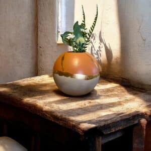 Vase rond beige et terracotta avec un liseré en feuille d’or posé sur une table en bois rustique avec un petit bouquet