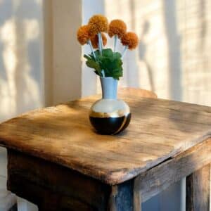 Vase étroit et rond en bas noir et gris avec un liseré en feuille d’or posée sur une table en bois rustique Avec un bouquet de fleurs orange