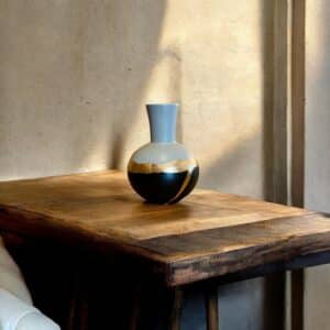 Vase étroit et rond en bas noir et gris avec un liseré en feuille d’or posée sur une table en bois rustique