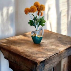 Petit vase gris et vert avec un liseré en feuille d’or, avec deux anses, posé sur une table en bois rustique, avec un bouquet de fleurs orange