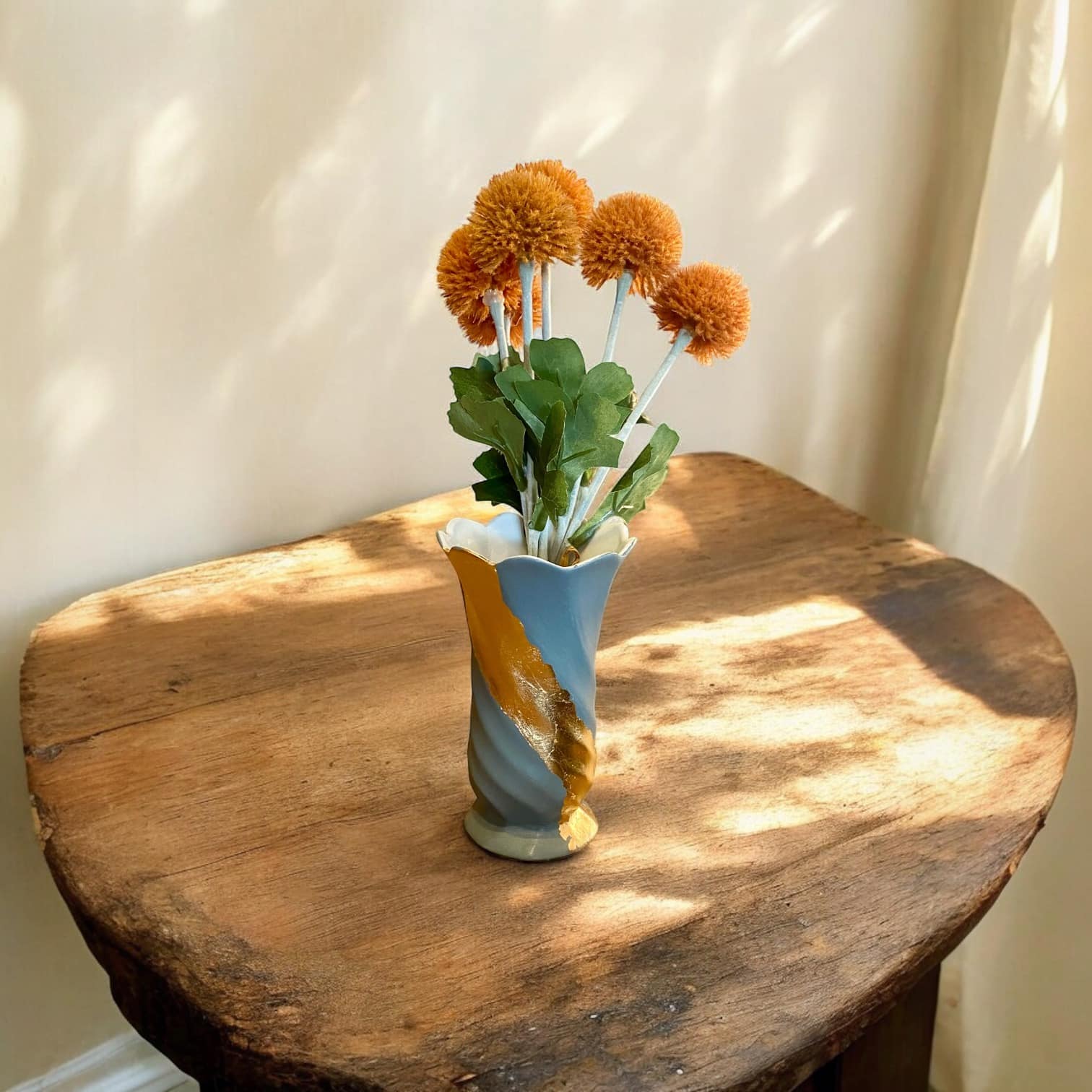 Petit vase gris bleu torsadé avec un liseré en feuille d’or posé sur une table en bois rustique Avec un bouquet de fleurs orange
