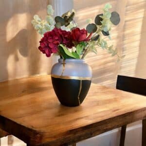 Grand vase noir et gris avec un liseré en feuille d’or avec un bouquet de fleurs rouges posé sur une table en bois rustique