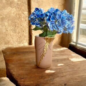 Grand pichet rose poudré avec moulure en feuille en liseré en feuille d’or posé sur une table en bois rustique avec un bouquet de fleurs bleues