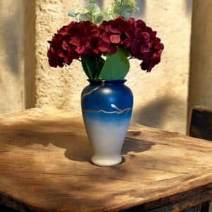 Vase dégradé bleu tirant sur ke beige avec un liseré fin en feuille d’or posé sur une table en bois rustique, avec un bouquet de grosses fleurs rouges