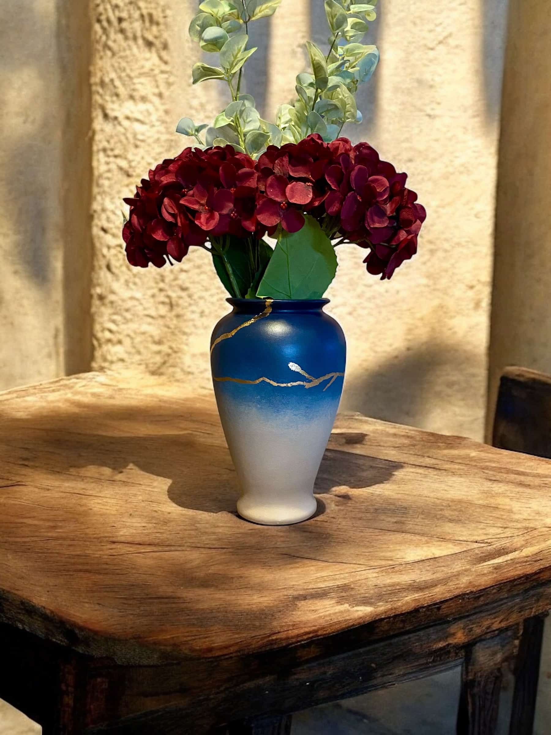 Vase dégradé bleu tirant sur ke beige avec un liseré fin en feuille d’or posé sur une table en bois rustique, avec un bouquet de grosses fleurs rouges