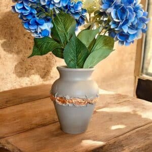 Vase en terre cuite relooké en beige avec une moulure et de la feuille d’or couleur bronze, avec un bouquet de fleurs bleues, posé sur une table rustique