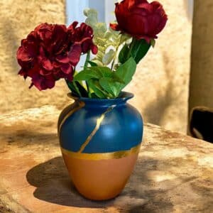 Grand vase terracotta et bleu marine avec un liseré en feuille d’or posé sur une table en bois rustique, avec un bouquet de fleurs rouges