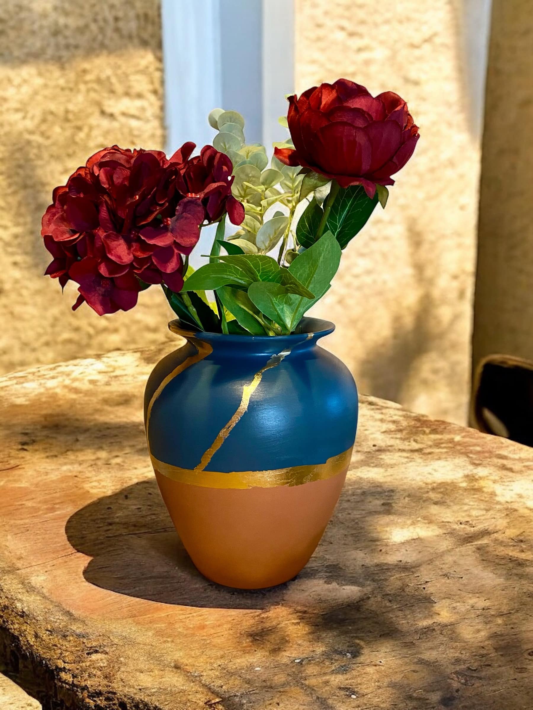 Grand vase terracotta et bleu marine avec un liseré en feuille d’or posé sur une table en bois rustique, avec un bouquet de fleurs rouges